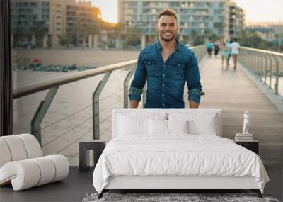 A young handsome man on the bridge near beautiful beach. Rich and successful male model wearing denim shirt Wall mural