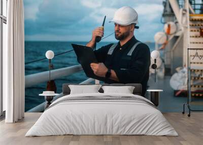 Marine Deck Officer or Chief mate on deck of offshore vessel or ship , wearing PPE personal protective equipment - helmet, coverall. He holds VHF walkie-talkie radio in hands. Wall mural