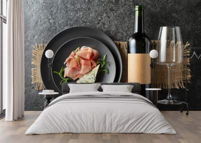 Bottle of red wine with blue cheese, prosciutto, and rosemary on a black table. Wall mural