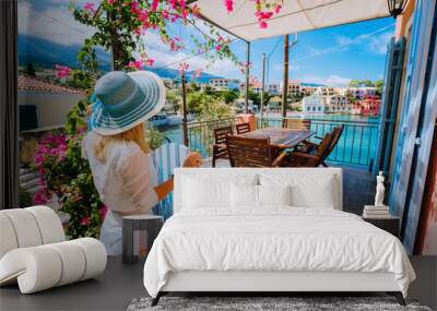 Female tourist with blue hat staying in Assos village in front of cozy veranda and admiring turquoise colored bay of Mediterranean sea and beautiful colorful houses of in Kefalonia, Greece Wall mural