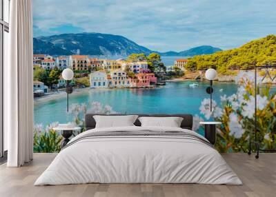 Panoramic view to Assos village in Kefalonia, Greece. Bright white blossom flower in foreground of turquoise colored calm bay of Mediterranean sea and beautiful colorful houses in background Wall mural