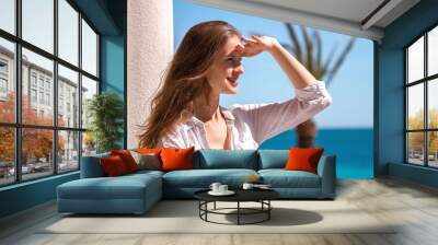 Beautiful smiling woman looking into the distance covering her eyes with the palm of the sun on background of blue sea and sky. Wall mural