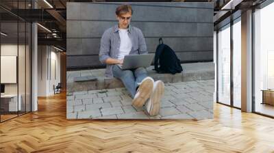 Young teenage boy student in casual backpack glasses holding laptop computer isolated on wall. Man working on laptop. The concept of studying at a university college Wall mural