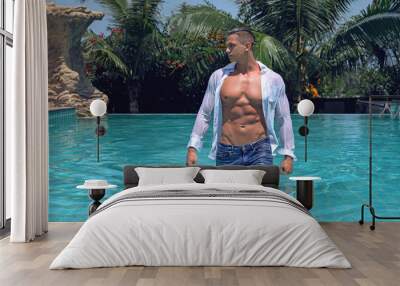 Muscular handsome sexy young wet man with athletic body is standing in the pool in blue jeans and white shirt. Fitness male model posing on camera Wall mural