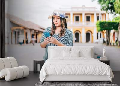 Beautiful young tourist woman in hat using phone on the street. Latin tourist girl texting with phone on the street Wall mural