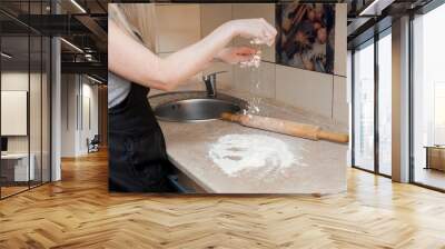 A woman prepares food from flour. On the table are kitchen wooden utensils for making dough. Girl sprinkles flour on the table. A live shot of a woman's everyday life at home. Wall mural