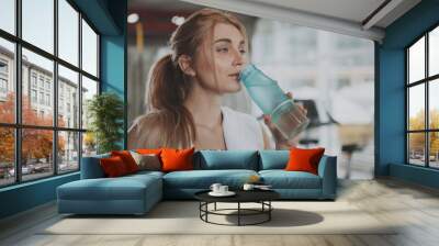 Close up of a gorgeous young sportswoman looking away, drinking water after exercising. Charming healthy blond haired female athlete resting at the gym after working out, copy space Wall mural