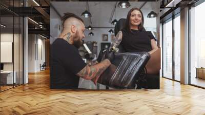 Low angle shot of a beautiful young woman getting arm sleeve tattoo by professional tattooer. Tattoo artist working at his salon, tattooing lovely female customer Wall mural