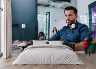 Camera view of gorgeous Caucasian man in shirt working in business company. Busy specialist looking directly at screen and using keyboard to type message. In background people sitting at computers. Wall mural