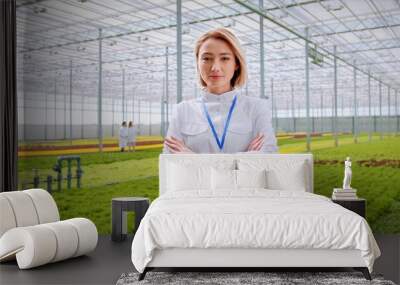 Caucasian female biologist in white lab coat posing in modern hydroponic farm with crossed arms. Confident woman with blond hair testing national standards of organic vegetables. Wall mural