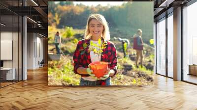 Portrait shot of blonde young beautiful Caucasian woman standing outdoor with tree seedling in pot and smiling cheerfully. Pretty female eco activist working against deforestation. Gardening. Wall mural