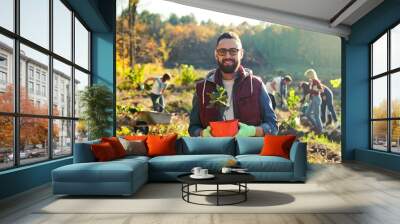 Portrait shot of young handsome Caucasian man in glasses standing outside with tree seedling in pot and smiling cheerfully. Male eco activist working against deforestation. Gardening concept. Wall mural