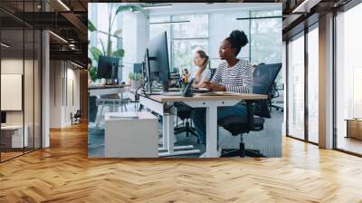 Zooming in on pretty woman with casual clothes working at computer in office. Distracted by camera from work and joyfully smiling. In background people sitting at their workplaces. Job concept. Wall mural