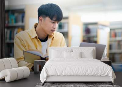 A young man is sitting at a table in a library, reading a book Wall mural