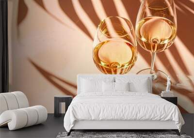 Two glasses of white wine in pale pink tinted glass featuring palm leaf shadows cast by sunlight embody a creative summer drink minimal concept They are set against a peach colored background with am Wall mural