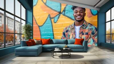 A young man stands confidently in front of a colorful graffiti mural, showcasing a joyful smile and casual attire in a lively urban environment Wall mural