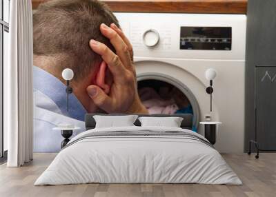 A man holds his head and is worried about the problem of washing clothes. the washing machine breaks or the laundry deteriorates when washing in the washing machine. Wall mural