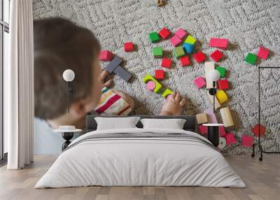 A little boy, 2 years old, sits on the floor and carpet at home and plays with colored wooden blocks. Educational toy constructor. The child learns to play according to the Montessori method. Top view Wall mural