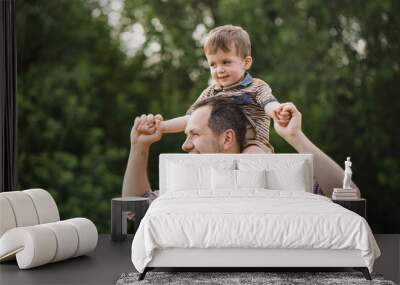 father rolls his little son on his shoulders. dad and child play and laugh in the garden on a warm summer day. a bearded man in a plaid shirt and a little boy roll around his neck Wall mural