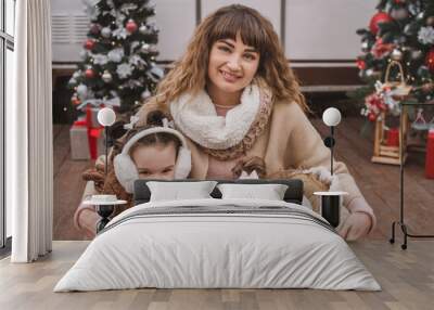 Mom and two twin daughters dressed in stylish fur coats and winter fur headphones play, hug and enjoy near the trailer in the forest with New Year's decorations. Christmas atmosphere and comfort Wall mural