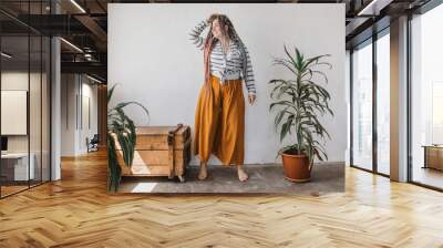 Positive smiling plus size woman with dreadlocks in bright yellow pants in a modern bohemian boho interior is meditating surrounded by large flowers and plants. Ecological approach to conscious consum Wall mural