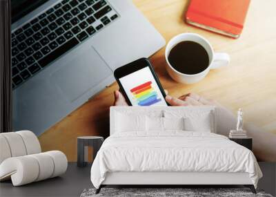 A mobile phone with a painted rainbow flag on the screen in the hands of a person near a laptop keyboard. LGBT concept and technology concept Wall mural