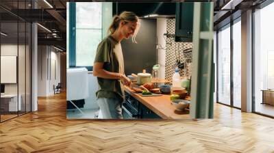 A young happy blond smiling woman cooking food in a home kitchen.. Wall mural