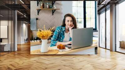 Business thirty professional woman working on laptop in the kitchen at home. A real female freelancer uses a computer to work remotely from home. Wall mural