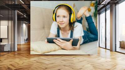 Nice redhead girl child lying on the couch with an electronic tablet listens to music in yellow headphones. Remote learning, teenager using wireless devices for education. Wall mural