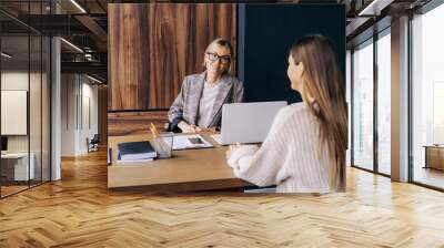 Two young modern business women smiling talking while sitting at the desk in the office. Wall mural