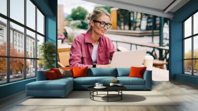 Young blonde business woman sitting on the terrace in a cafe working on a laptop. Wall mural