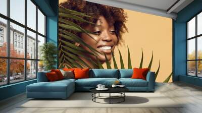 cheerful African American woman with a healthy clean skin near palm leaves on a beige background with a free copy space. Smiling african american girl having afro style hair among tropical leaf. Wall mural