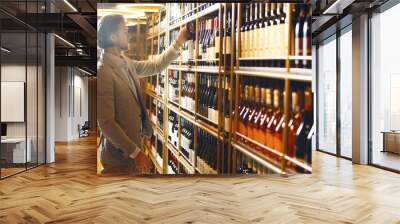 Sommelier stands in cellar and taking out one wine of many submitted from wooden shelf. Male bartender in wine storage choose necessary alcoholic bottle for buyer based on his requests. Wall mural