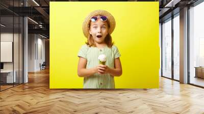 Studio portrait of little girl in summer dress, expresses surprise emotion, wears straw hat and sunglasses, holding ice cream on hand, stands over yellow isolated. Wall mural