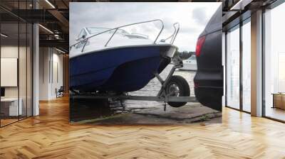 A 4x4 black car pulls blue-white trimaran cabin motor boat on trailer on concrete slipway into the river water, boat launch before active recreation on the river Wall mural