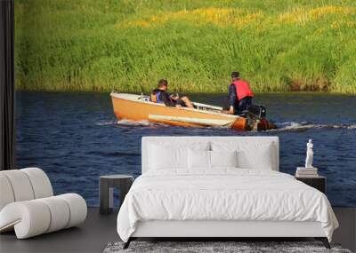 Two men in life jackets floating in a small orange fishing outboard motor boat along the shore with green grass, rear side view Wall mural