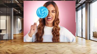 Portrait, funny face and lollipop with a woman in studio on a pink background holding giant candy. Comic, comedy and sweet with a playful young female feeling silly while eating a blue sugar treat Wall mural