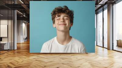 Portrait of a young smiling cheerful boy student wearing casual white t-shirt standing with closed eyes isolated on a studio blue background. People emotions and lifestyle, Generative AI Wall mural