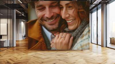 A close-up image of a happy couple, newly engaged, focusing on the woman's hand with a sparkling diamond ring. They are smiling and embracing each other outdoors Wall mural