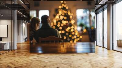 A man and a woman are seated at a table in front of a decorated Christmas tree, enjoying a festive moment together Wall mural