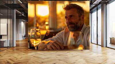 A man with a beard and glasses is sitting at a table with a glass of wine. He is looking at the camera with a smile on his face Wall mural