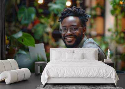 A man with glasses and a beard is smiling and holding a piece of paper. He is sitting in a green room with plants around him Wall mural