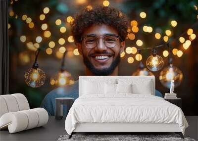 A man with glasses and a beard is smiling in front of a string of lights. The lights are glowing and creating a warm, inviting atmosphere Wall mural