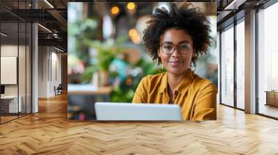 A woman with curly hair and glasses is sitting at a table with a laptop. She is smiling and she is enjoying her time Wall mural