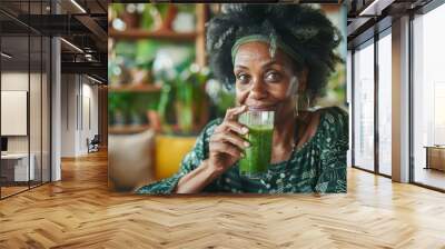 A woman with dark hair and a green headband sits in a cafe, smiling and holding a glass of green smoothie Wall mural