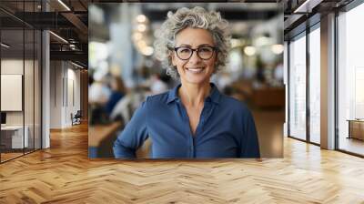 A woman with gray curly hair and glasses smiles confidently at the camera while standing in a modern office setting Wall mural