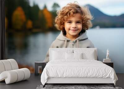 A young boy with curly hair is smiling and wearing a grey hoodie. He is standing by a body of water, possibly a lake or a river. Concept of warmth and happiness Wall mural