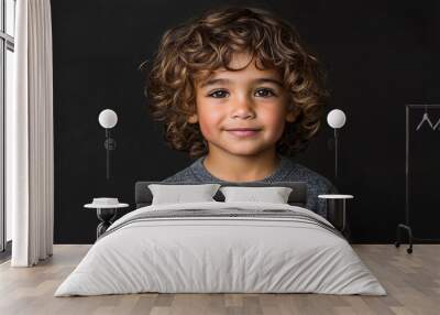 A young boy with curly hair is smiling at the camera. He is wearing a gray sweater and is standing in front of a black background Wall mural