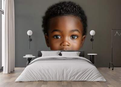 A young child with dark skin and curly black hair looks directly at the camera. The child is wearing a white shirt and is positioned in front of a gray background Wall mural