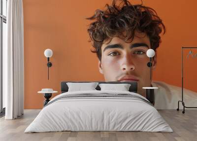 A young man with curly brown hair stares directly at the camera, standing against a bright orange wall. He is wearing a plain white t-shirt Wall mural